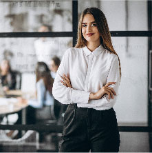 Mujer vestida formal ejecutiva con los brazos cruzados sonriendo y al fondo un grupo de personas en una oficina.