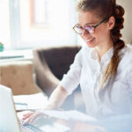 Mujer joven y sonriente, en oficina iluminada revisando su laptop.