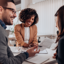 Tres personas jóvenes, dos mujeres y un hombre sentados alrededor de un escritorio con documentos compartiendo y charlando sonriendo.