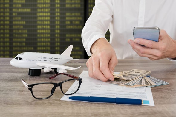 Sobre una superficie hay un documento, una persona sosteniendo varios billetes, unos lentes de ver, un lapicero y un avión pequeño
