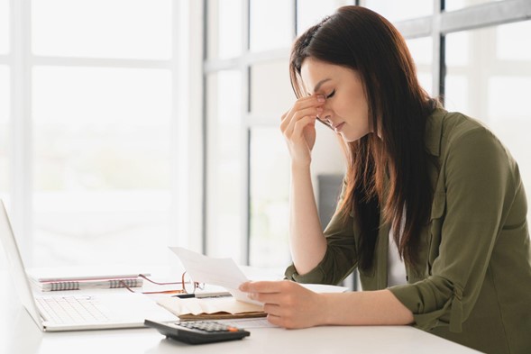 Mujer con cara de preocupación, apoyada en una mesa donde se encuentra una computadora, varios documentos y una calculadora