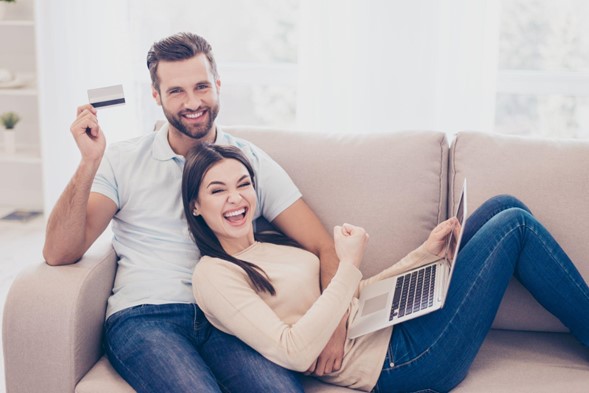 una pareja acomodada en el sillón, con una laptop en las piernas, mostrando una tarjeta y con cara de felicidad en ambas personas