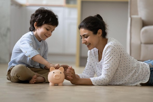 Mujer acostada en el piso en compañía de un niño, ambos echando monedas a un chancho de cerámica.