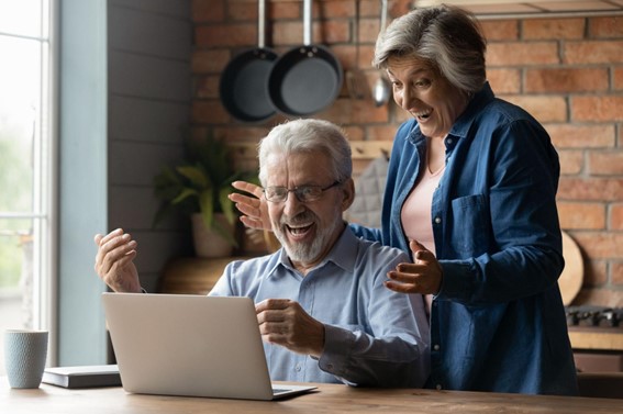 una pareja de adultos mayores felices, viendo una computadora que tienen al frente, sobre una mesa