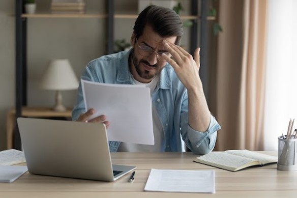 Hombre con cara de preocupación, sentado en un escritorio, con documentos en la mano y con la computadora al frente.