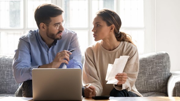 Dos personas hablando y sentadas en un sillón, con una laptop al frente y con documentos en la mano.