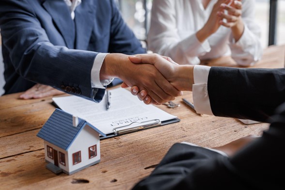 2 personas dándose la mano sobre una mesa, cerrando un trato y con una casa pequeña y documentos, decorando la mesa.