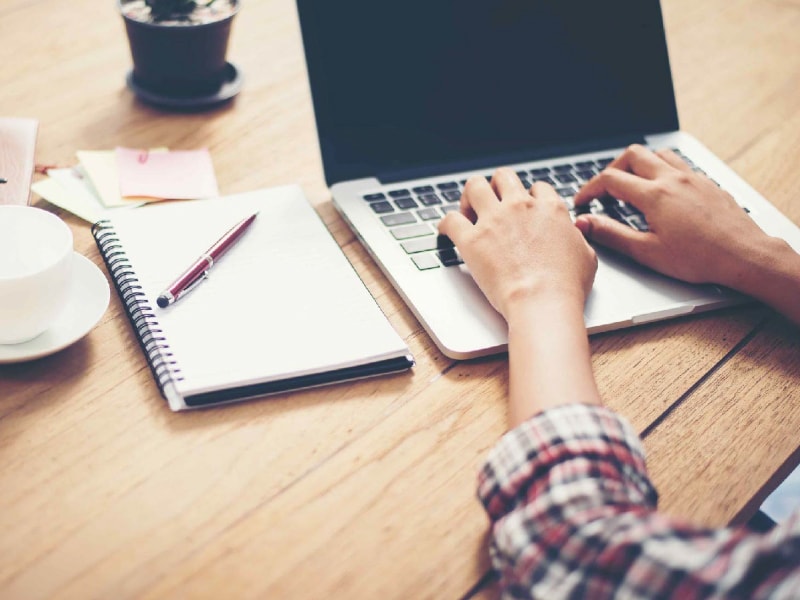 Persona digitando en su laptop en un escritorio junto a una libreta con apuntes.