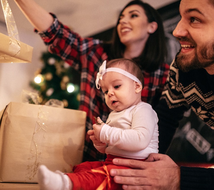 Familia de tres (padre, madre, hija) con suéteres blancos y gorros de Santa, sosteniendo bengalas encendidas junto al árbol