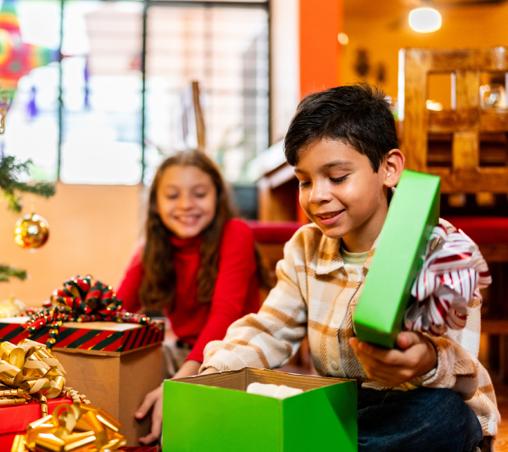 Dos niños sentados en el suelo, el niño sonríe al abrir una caja de regalo verde junto a regalos apilados y un arbol de Navidad con adornos dorados y rojos.