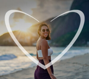 Mujer joven sonriendo y con anteojos de sol mira a cámara, detrás de ella un atardecer cae en el mar y a la par de una colina. En la fotografía, hay un corazón blanco superpuesto.