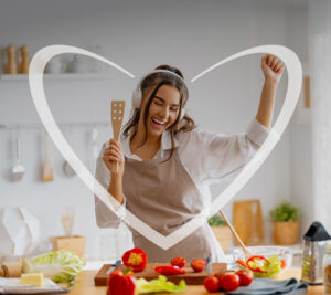 Mujer joven sonriendo en su cocina mientras prepara una ensalada fresca, mostrando un momento de alimentación saludable y bienestar. En la imagen, hay un corazón blanco superpuesto.
