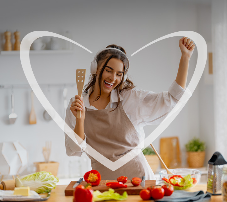 Mujer joven sonriendo en su cocina mientras prepara una ensalada fresca, mostrando un momento de alimentación saludable y bienestar. En la imagen, hay un corazón blanco superpuesto.