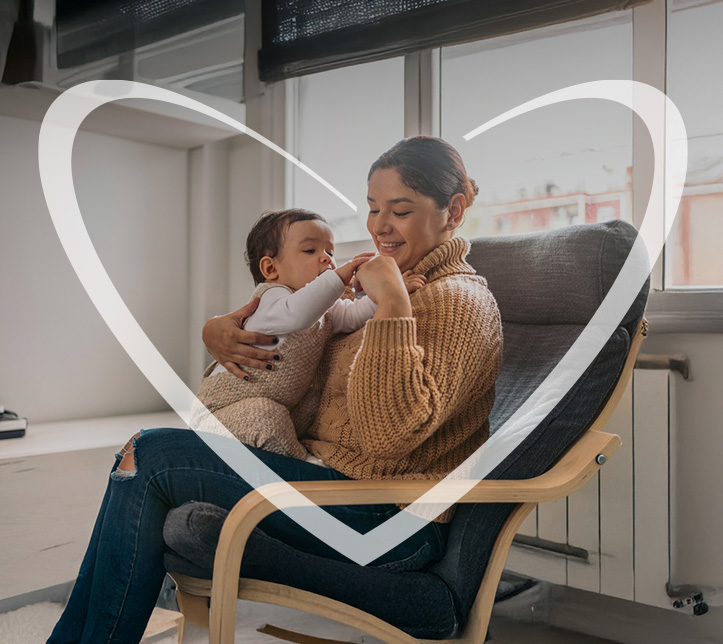 Crianza consciente de una Mujer joven sonriendo mientras sostiene a su bebé en brazos, sentada en la sala iluminada, transmitiendo calma, conexión y ternura. En la imagen, hay un corazón blanco superpuesto.