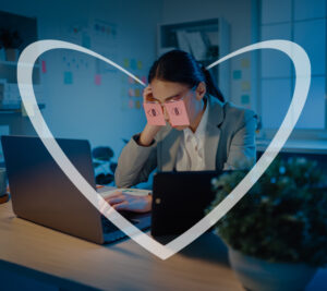 Una empresaria joven está frente a su computadora con dos post-its pegados sobre los ojos, cada uno con ojos dibujados, intentando aparentar que sigue despierta y trabajando. En realidad, parece agotada y dormida frente a la pantalla. En la imagen, hay un corazón blanco superpuesto.