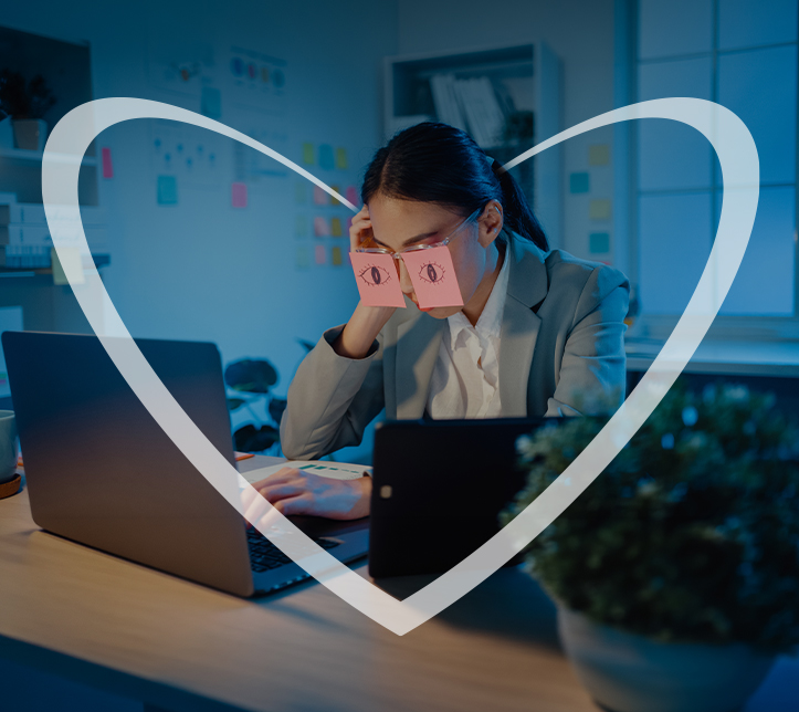 Una empresaria joven está frente a su computadora con dos post-its pegados sobre los ojos, cada uno con ojos dibujados, intentando aparentar que sigue despierta y trabajando. En realidad, parece agotada y dormida frente a la pantalla. En la imagen, hay un corazón blanco superpuesto.