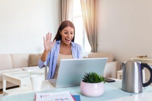 Mujer muy feliz sentada en la mesa de su casa, saludando a la pantalla de la computadora que tiene al frente.