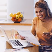 imagen de mujer con un papel en la mano y un celular, sentada en una silla y en la mesa hay una computadora