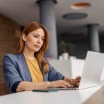 Portrait of smart casual female executive sitting at corporate office and typing on a laptop.