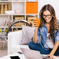 mujer sentada en un sillon con una tasa naranja en la mano y con la otra mano utilizando una computadora