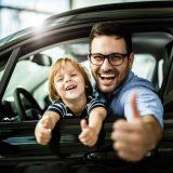 Happy single father and son testing new car in a showroom and showing thumbs up.